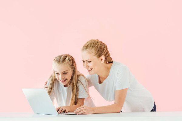 smiling mother and daughter in white t-shirts using laptop together isolated on pink