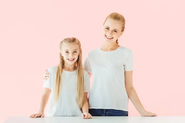 beautiful mother and daughter in white t-shirts looking at camera isolated on pink