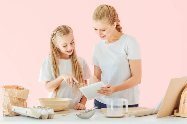 attractive mother and little daughter in white t-shirts using tablet while cooking isolated on pink