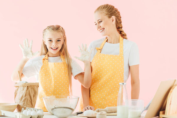 happy mother and daughter in yellow aprons cooking together isolated on pink