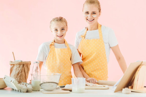 happy mother and daughter with flour on faces looking at camera while cooking isolated on pink