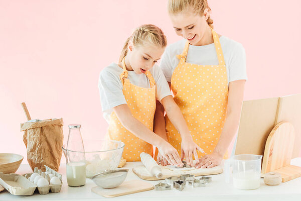 smiling mother and daughter cutting dough in different shapes for cookies isolated on pink