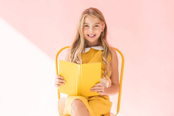 smiling fashionable kid reading book while sitting on yellow chair on pink
