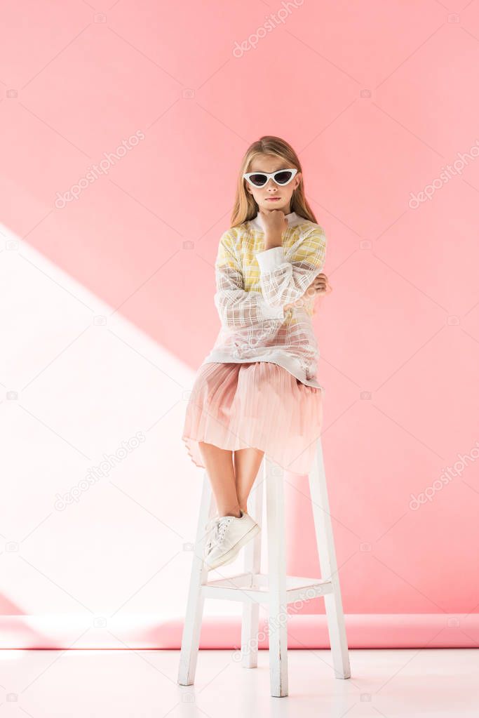 adorable pensive child in sunglasses sitting on stool on pink 