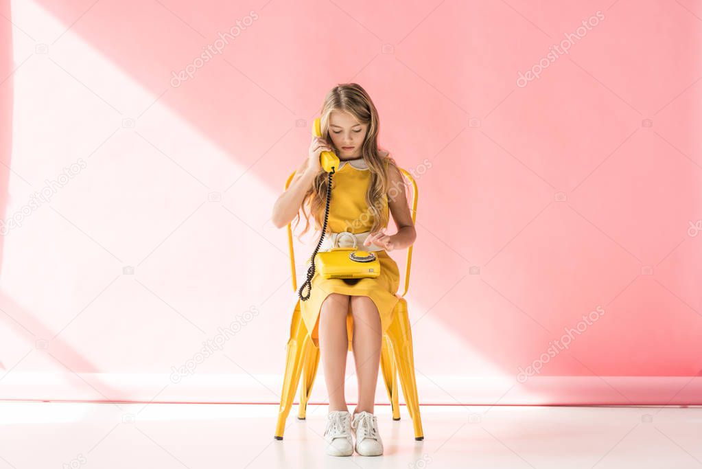 fashionable kid making call on retro phone while sitting on yellow chair on pink