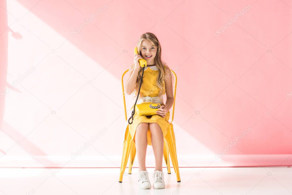 stylish smiling youngster talking on retro phone while sitting on yellow chair on pink 