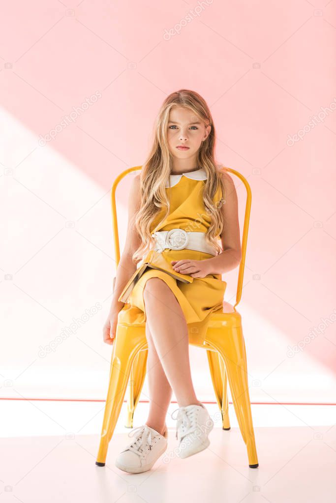 adorable blonde youngster with book sitting on yellow chair on pink