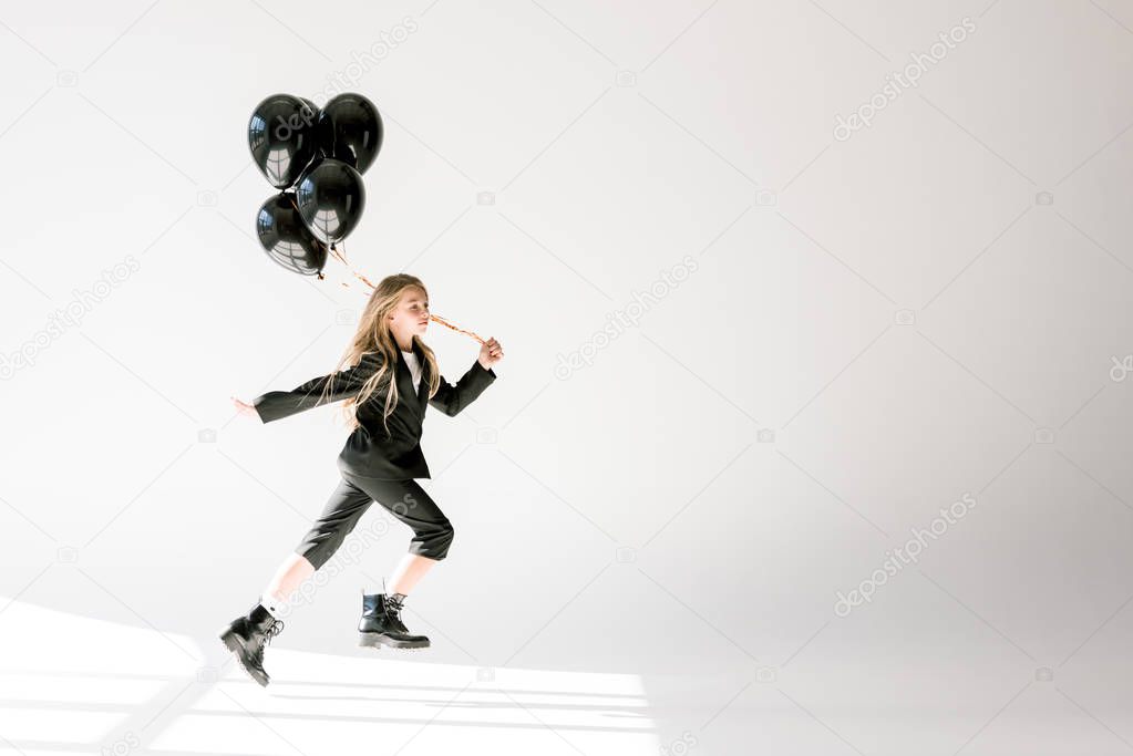 fashionable child in trendy suit jumping with black balloons on grey