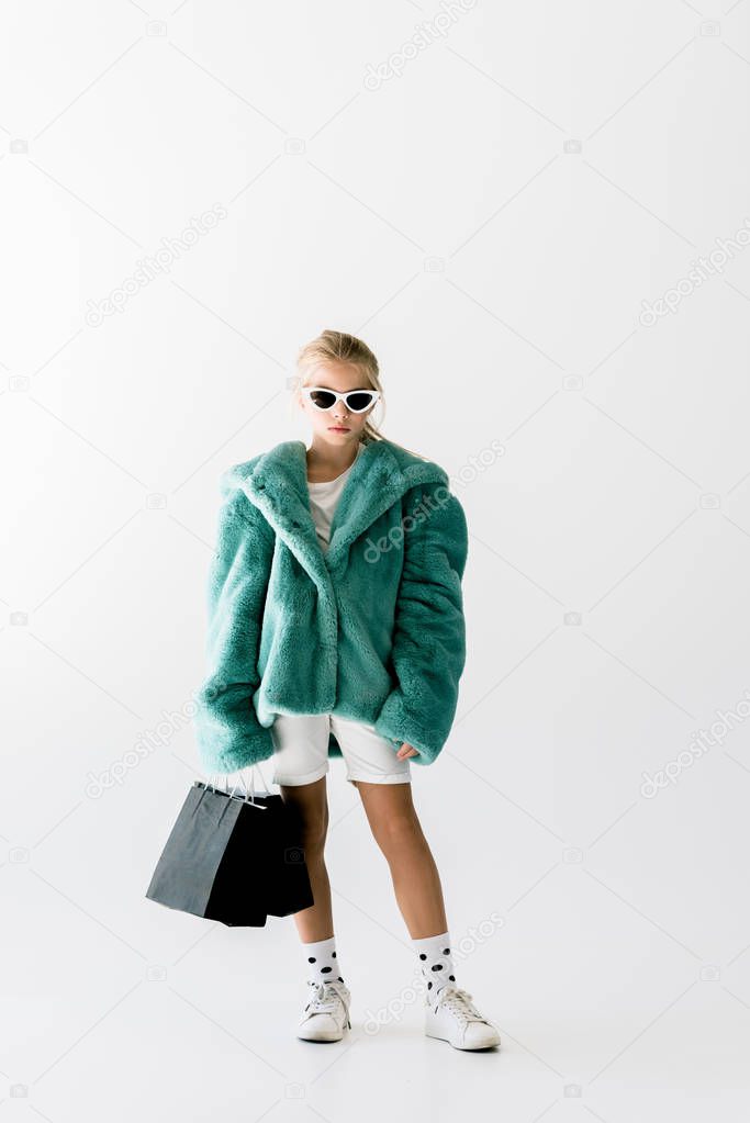 fashionable kid in turquoise fur coat posing with black shopping bags isolated on white