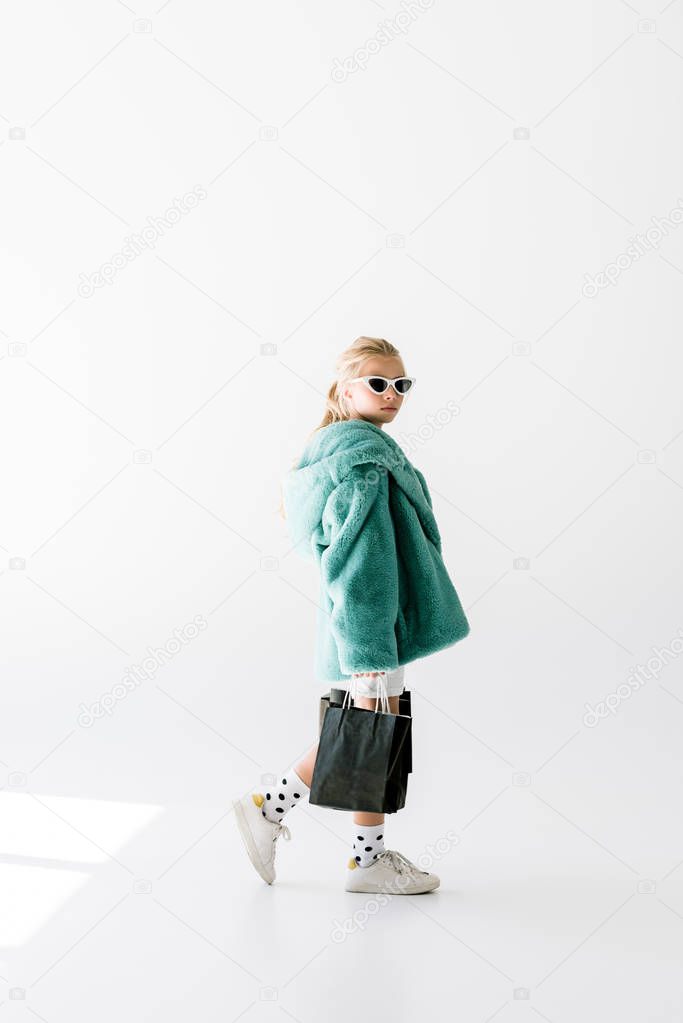 fashionable shopaholic in fur coat and sunglasses posing with black shopping bags on white
