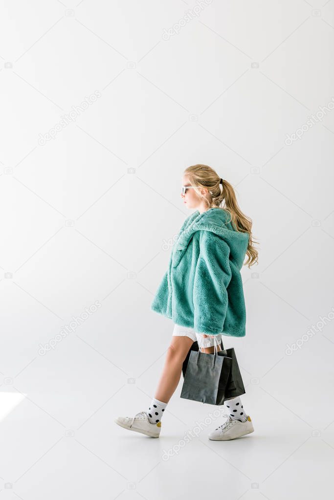 blonde female child in turquoise fur coat posing with black shopping bags on white