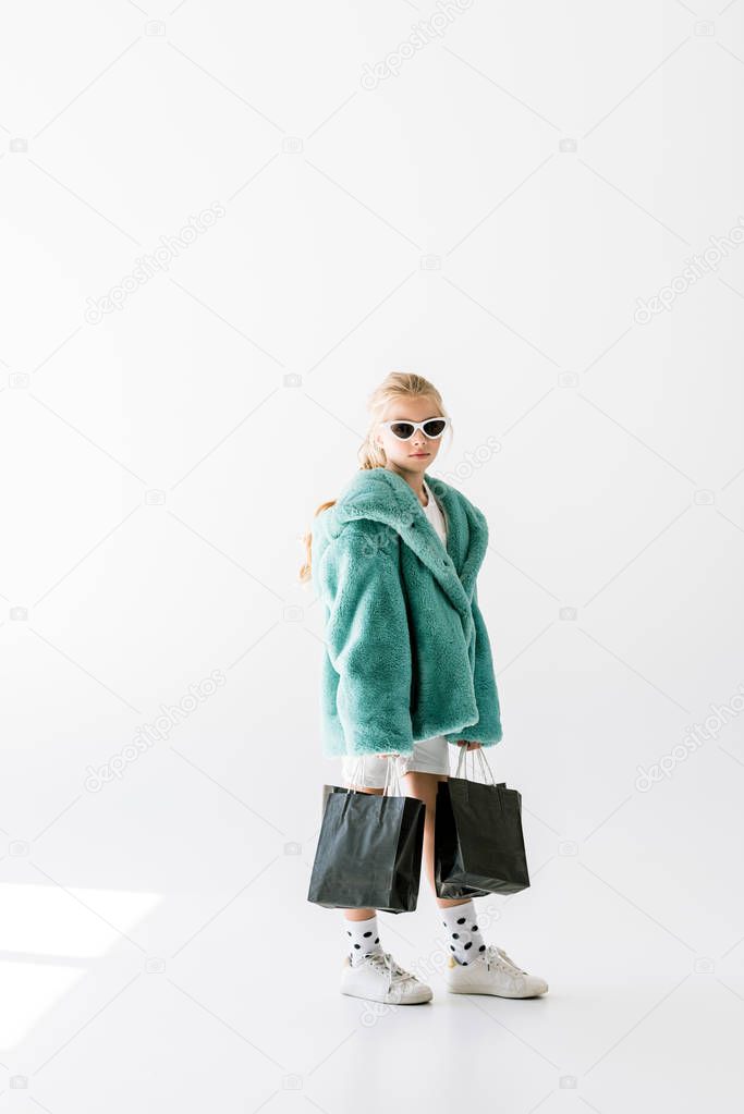charming kid in trendy fur coat and sunglasses posing with black shopping bags on white