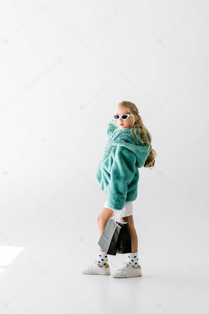 elegant child in turquoise fur coat and sunglasses posing with black shopping bags on white