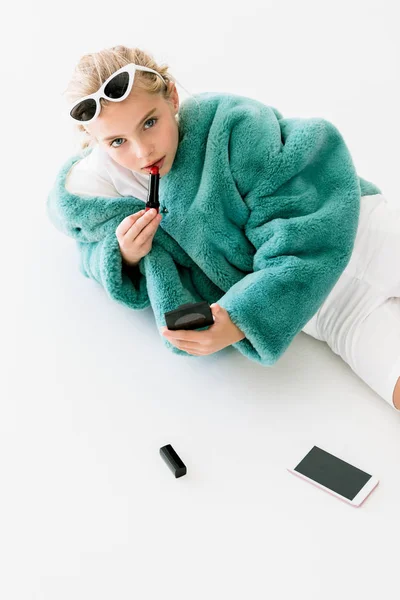 スマート フォンでホワイトに横たわっている間ミラーに口紅を適用する青緑色の毛皮のコートでスタイリッシュな金髪の子供 — ストック写真