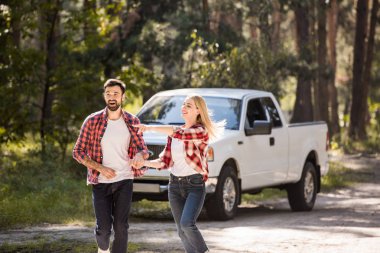 excited couple holding hands and running from pickup truck, woman showing something in forest clipart
