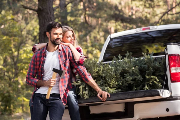 Couple with axe sitting on pickup truck with fir tree for christmas in forest