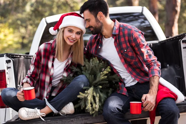 beautiful couple in santa hats holding red cups while sitting on pickup truck with fir tree for christmas