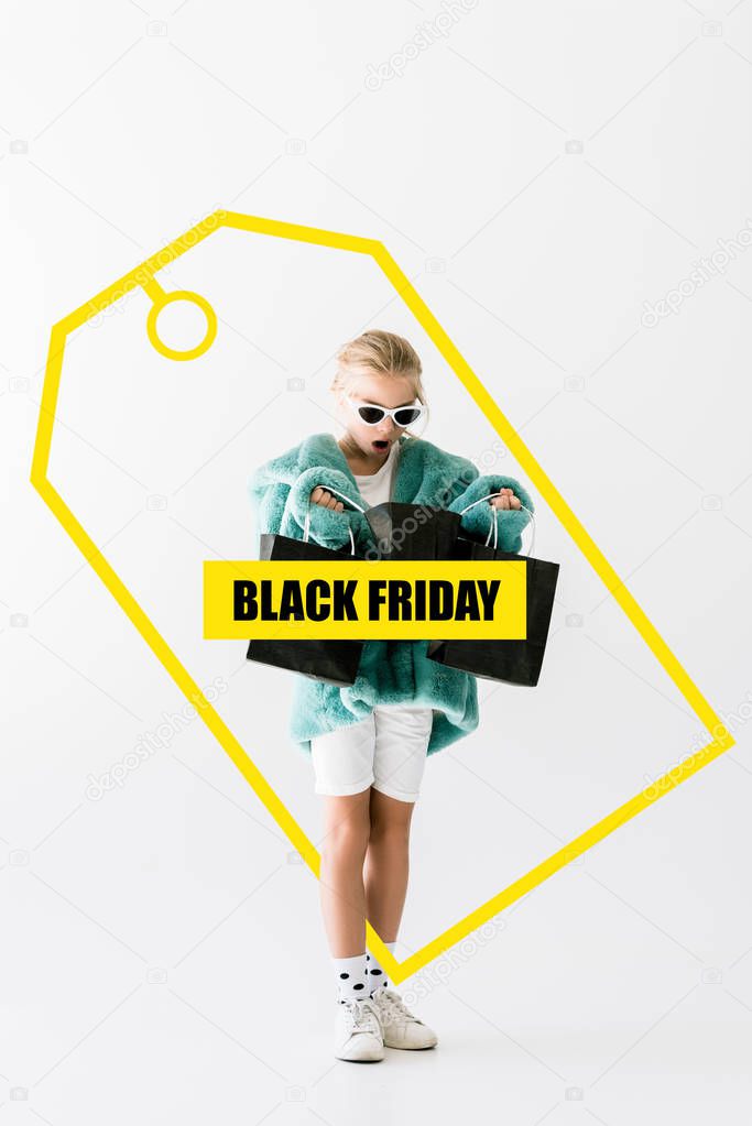 shocked kid in stylish fur coat looking into black shopping bags isolated on white, black friday sale banner concept