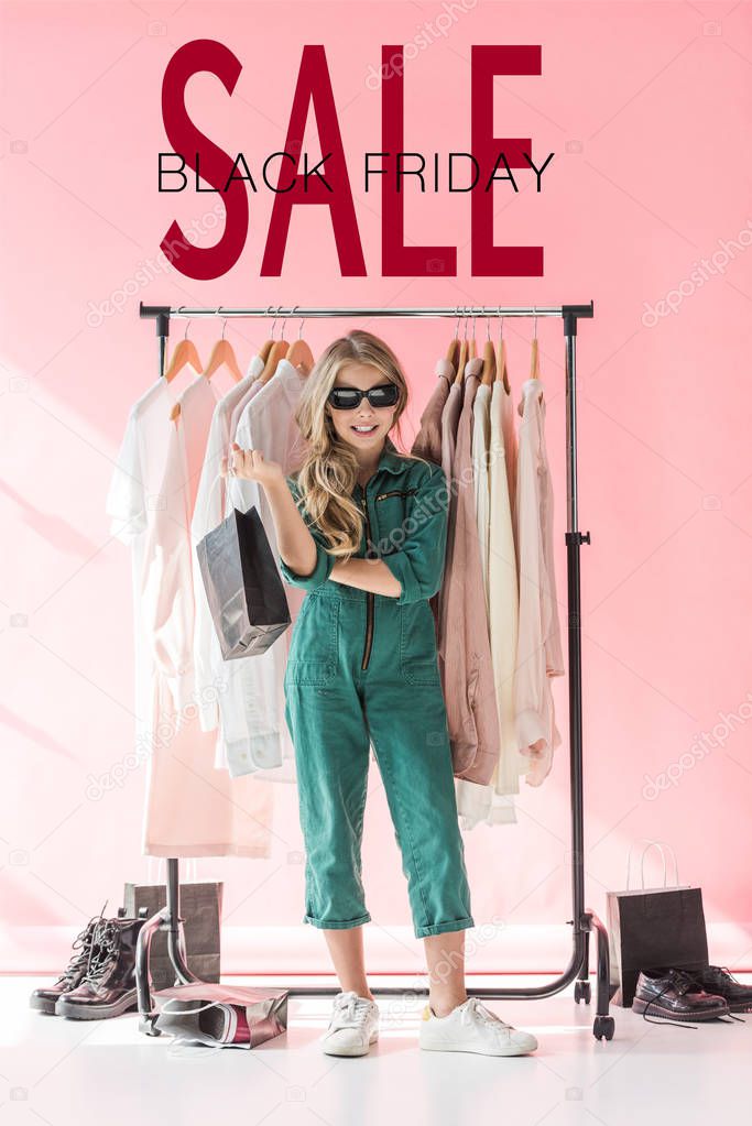 stylish child in overalls and sunglasses standing with shopping bag near clothes and footwear in boutique, black friday sale banner concept