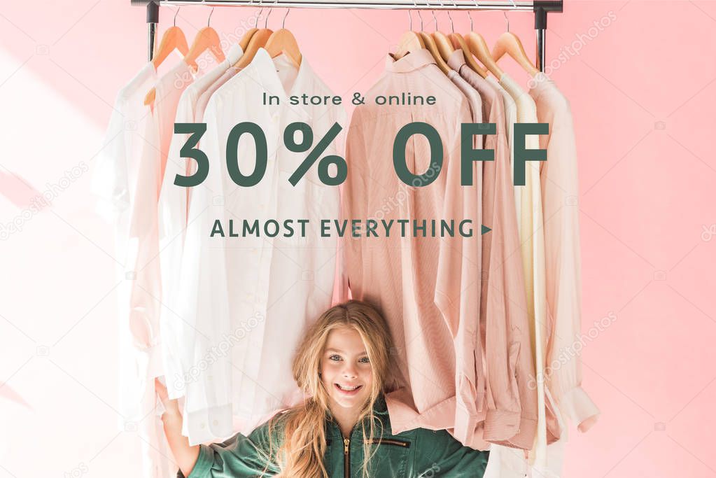 cheerful charming kid in trendy overalls sitting under clothes on hangers, store sale banner concept