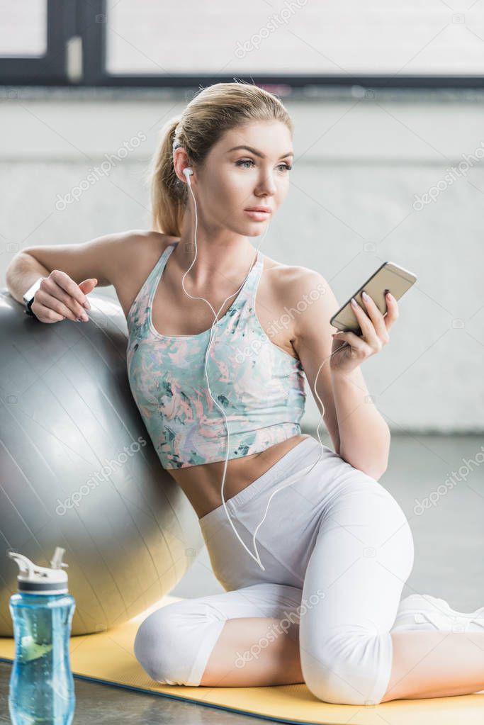 selective focus of female athlete in earphones resting and listening music with smartphone near fitness ball at gym