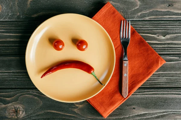 Sonriente triste hecha de pimienta y tomates en el plato con tenedor en la servilleta naranja - foto de stock