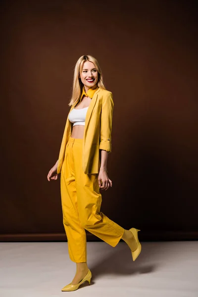 Volle Länge Ansicht der fröhlichen blonden Mädchen in stilvollem gelben Outfit lächelt und sieht weg auf braun — Stockfoto