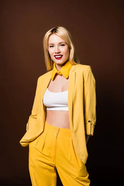 Belle jeune femme en vêtements jaunes élégants debout avec les mains dans les poches et souriant à la caméra isolée sur brun — Photo de stock