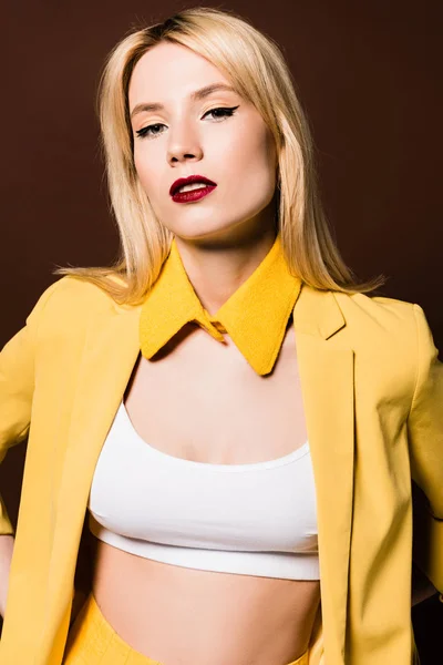 Portrait of beautiful blonde girl in fashionable yellow clothes looking at camera isolated on brown — Stock Photo