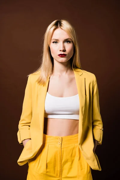 Porträt eines eleganten blonden Mädchens in gelben Kleidern, das mit den Händen in den Taschen steht und in die Kamera schaut, isoliert auf braun — Stockfoto