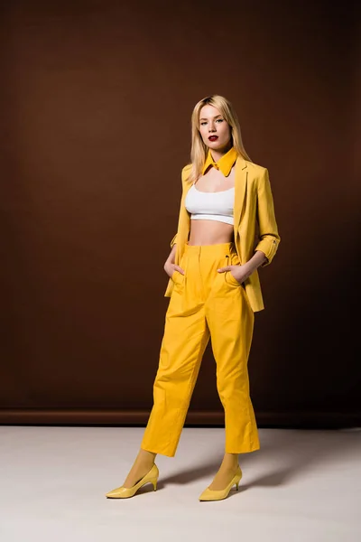 Schöne blonde Frau in stylischer gelber Kleidung, die Hände in den Taschen und Blick in die Kamera auf braun — Stockfoto