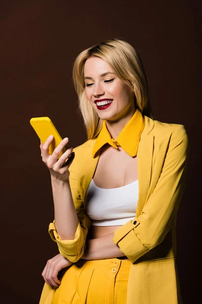 Feliz chica rubia con estilo utilizando teléfono inteligente amarillo aislado en marrón - foto de stock