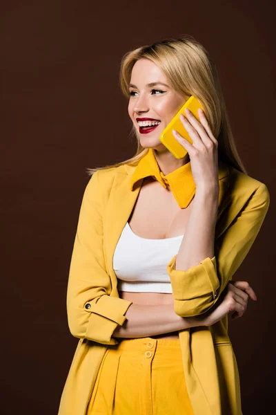 Alegre hermosa mujer rubia hablando por teléfono inteligente amarillo y mirando hacia otro lado aislado en marrón - foto de stock