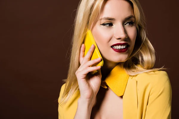 Hermosa chica rubia elegante hablando por teléfono inteligente amarillo y mirando hacia otro lado aislado en marrón - foto de stock