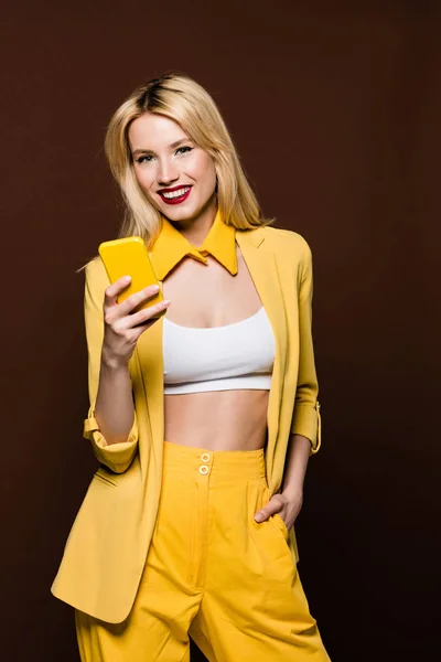 Hermosa mujer rubia elegante sosteniendo teléfono inteligente amarillo y sonriendo a la cámara aislada en marrón - foto de stock