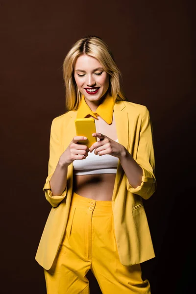 Menina loira elegante feliz usando smartphone amarelo isolado no marrom — Fotografia de Stock