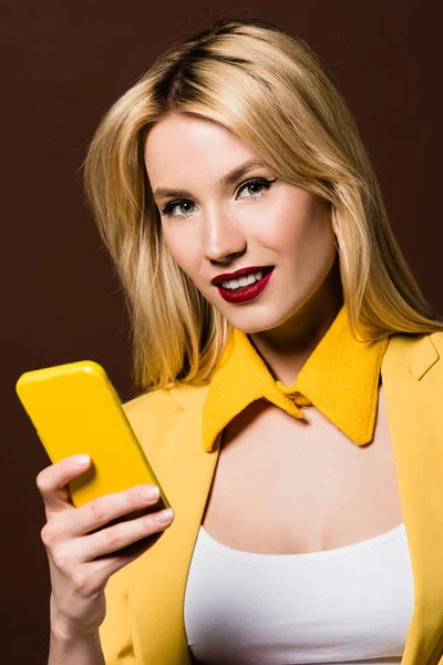 Beautiful stylish blonde girl using yellow smartphone and smiling at camera isolated on brown — Stock Photo