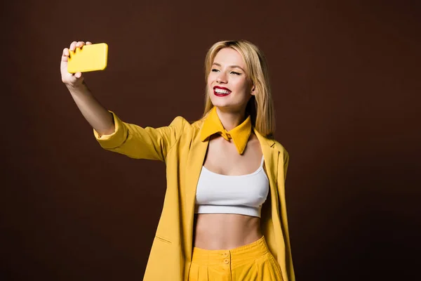 Belle fille blonde élégante souriante prenant selfie avec smartphone jaune isolé sur brun — Photo de stock