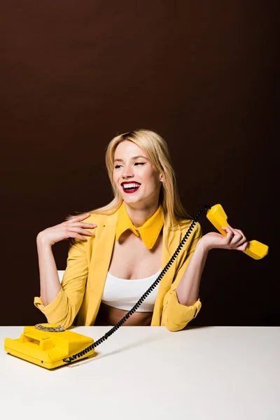 Hermosa chica rubia sonriente sosteniendo el auricular amarillo y mirando hacia otro lado en marrón - foto de stock