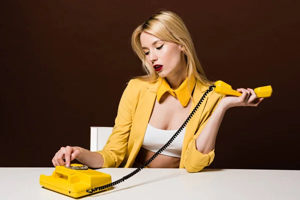 Beautiful stylish blonde woman in yellow clothes using rotary phone while sitting on brown — Stock Photo