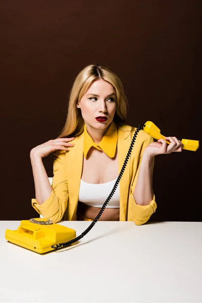 Atractiva chica rubia sosteniendo el auricular amarillo y mirando hacia otro lado en marrón - foto de stock