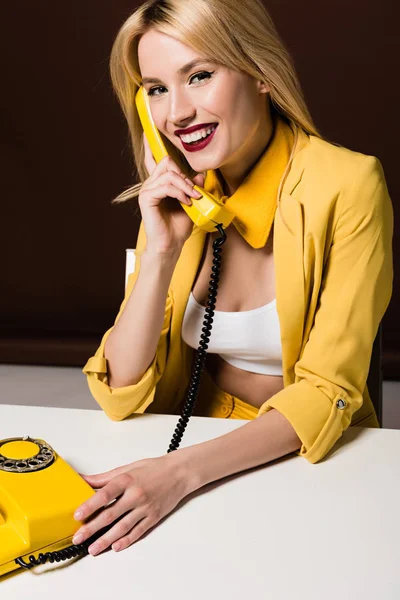 Hermosa chica rubia hablando por teléfono vintage amarillo y sonriendo a la cámara en marrón - foto de stock