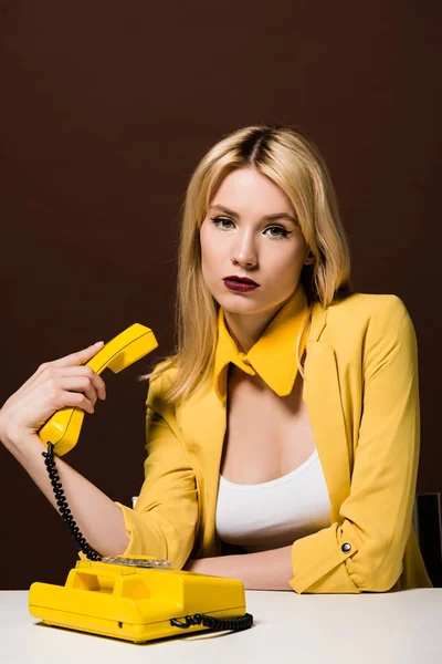 Hermosa chica rubia elegante sosteniendo el auricular amarillo y mirando a la cámara en marrón - foto de stock
