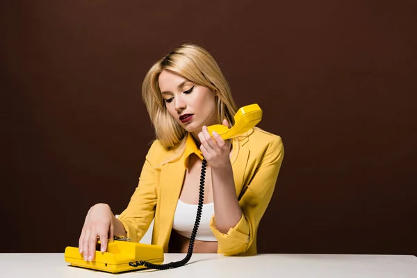 Chica rubia pensativa sosteniendo teléfono vintage amarillo mientras está sentado en marrón - foto de stock