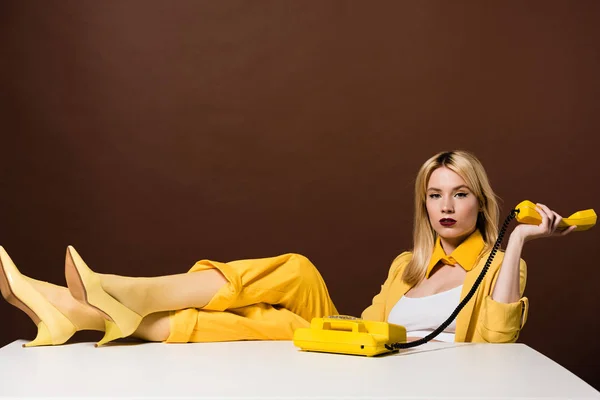 Bela menina loira elegante segurando aparelho amarelo e olhando para a câmera no marrom — Fotografia de Stock