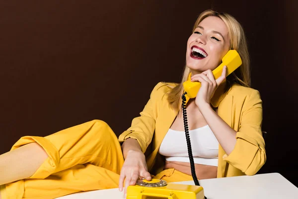 Весела блондинка розмовляє жовтим старовинним телефоном і сміється на коричневому — стокове фото
