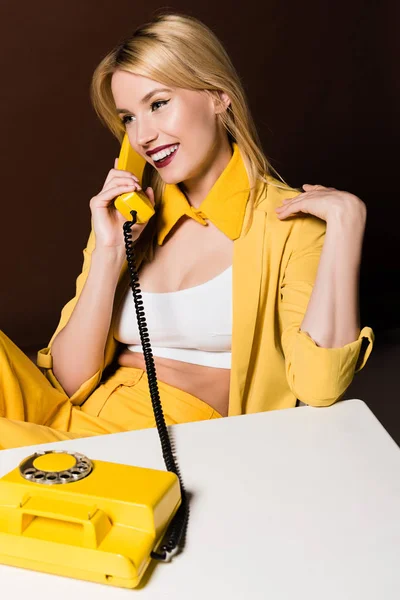 Schöne lächelnde stilvolle blonde Mädchen im Gespräch mit gelben Vintage-Telefon auf braun — Stockfoto