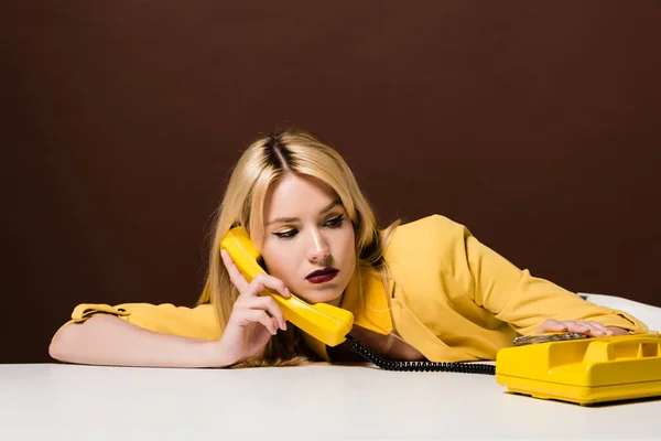 Attractive blonde girl in yellow clothes talking by vintage phone on brown — Stock Photo