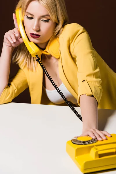 Beautiful pensive young woman talking by yellow rotary phone on brown — Stock Photo