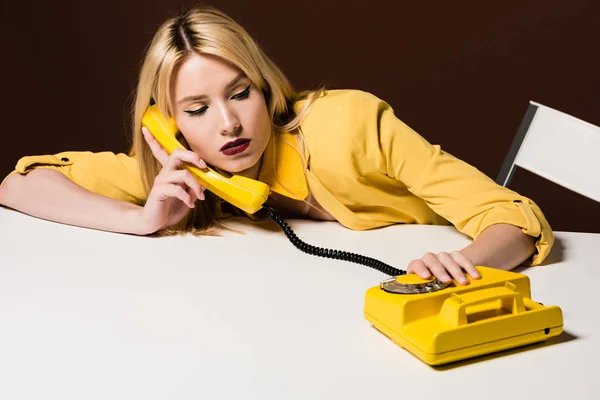 Красивая молодая женщина разговаривает по желтому роторному телефону на коричневом — стоковое фото